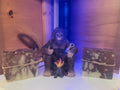 A central dark purple gorilla figurine seated at a small campfire, flanked by two rectangular marbled soap bars tied with twine, against a wood-paneled background with blue lighting.