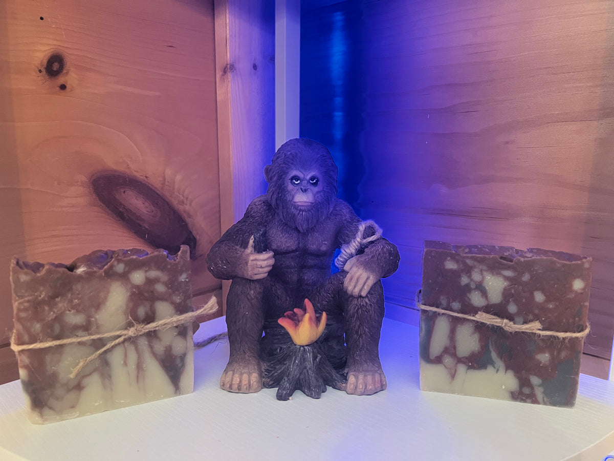 A central dark purple gorilla figurine seated at a small campfire, flanked by two rectangular marbled soap bars tied with twine, against a wood-paneled background with blue lighting.