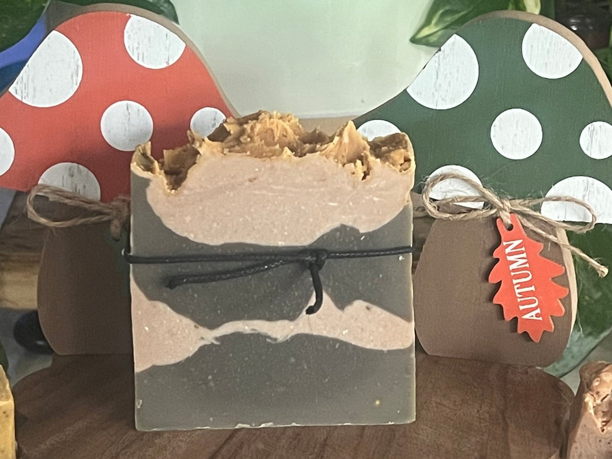 Three handmade artisanal soaps displayed on a wooden stand, styled with decorative mushroom-shaped signs and green houseplants in the background. The soaps are in earthy tones: one yellow bar, one layered beige and gray bar, and one speckled brown bar, each tied with twine.