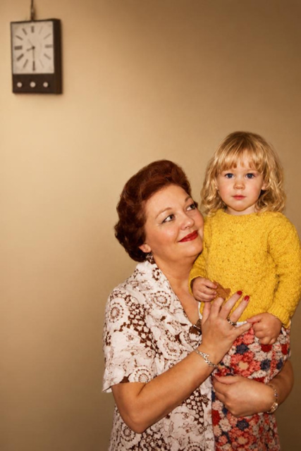 A woman holding a young child in a vintage-style indoor portrait. The woman has short reddish-brown hair and wears a cream lace-patterned blouse, while the child has blonde hair and wears a bright yellow knit sweater and a red floral skirt.